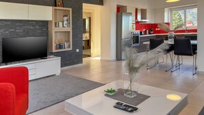 A modern living room with a flat-screen TV, a bookshelf, and a dining area with a kitchen in the background. - Villa Adam: Tranquility and Luxury with a Poolside View (Crete)