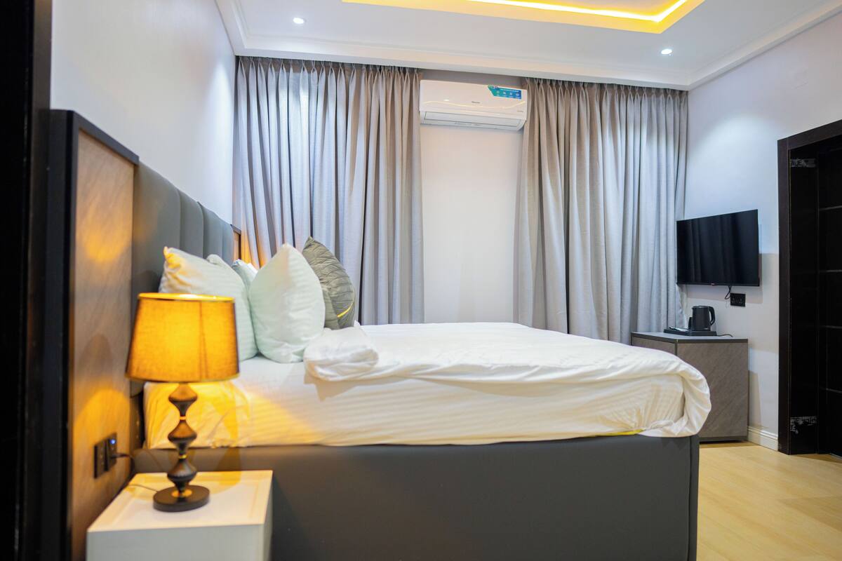 A modern hotel room with a neatly made bed, a bedside lamp, an air conditioning unit, and a television on a stand.