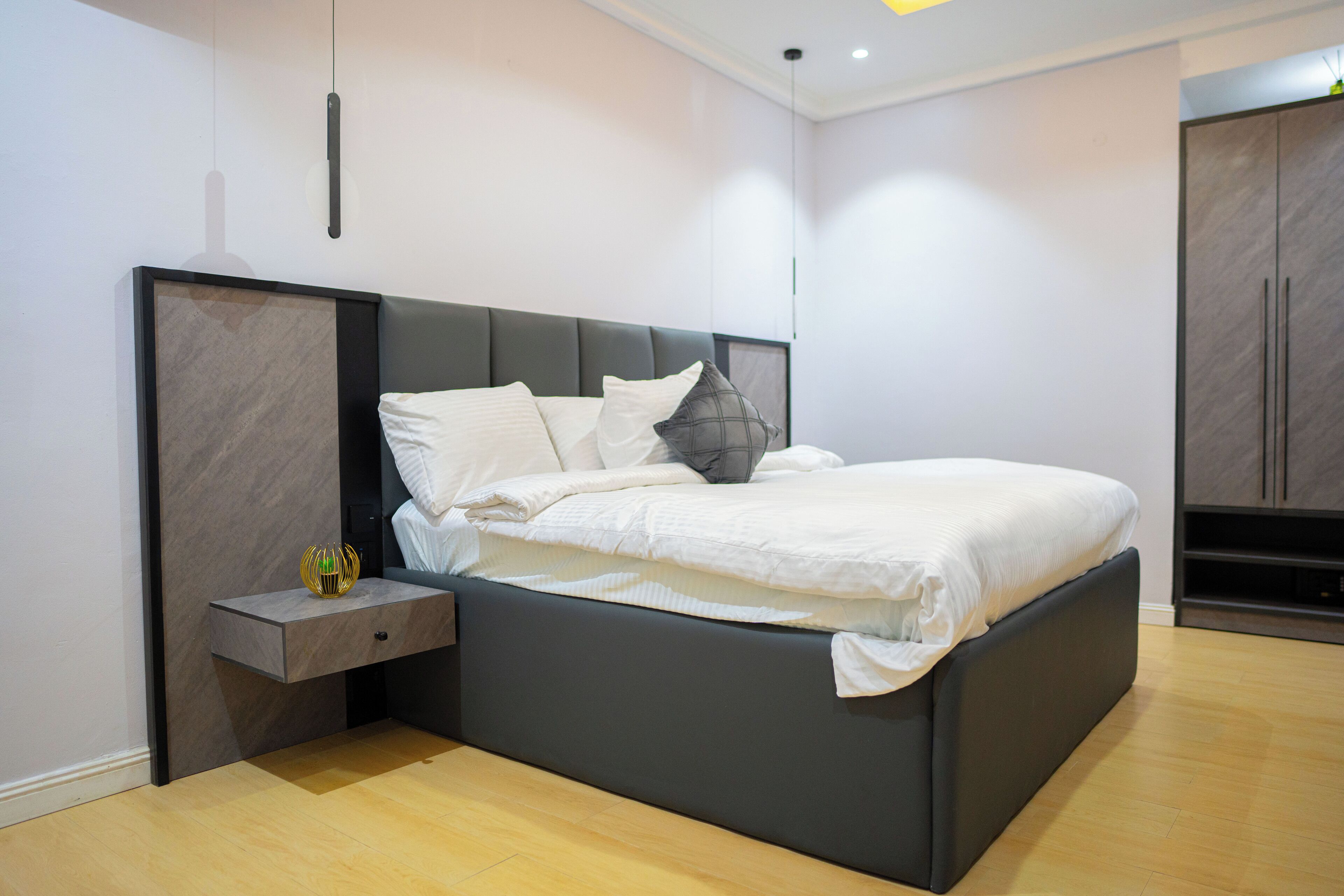A modern bedroom with a dark headboard, a bedside table with a lamp, and a wardrobe.