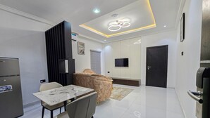 A modern living room with a dining area, a sofa, a TV mounted on the wall, and a black door. - Twinny Hotels & Suites (Lagos)