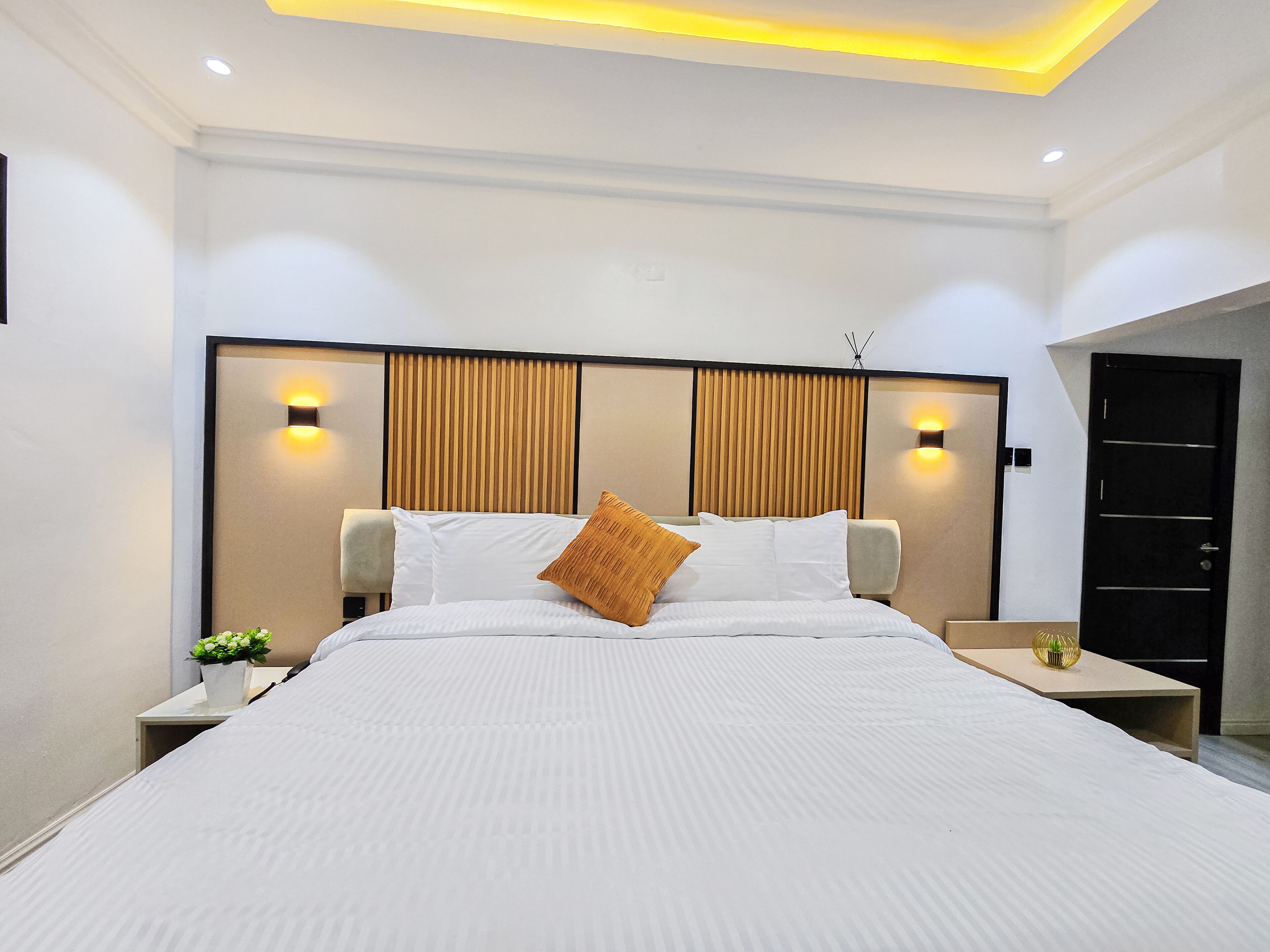 Ein modernes Hotelzimmer mit einem großen Bett, zwei Nachttischen, einer Ablagefläche und einer Wandleuchte sowie einer Tür.