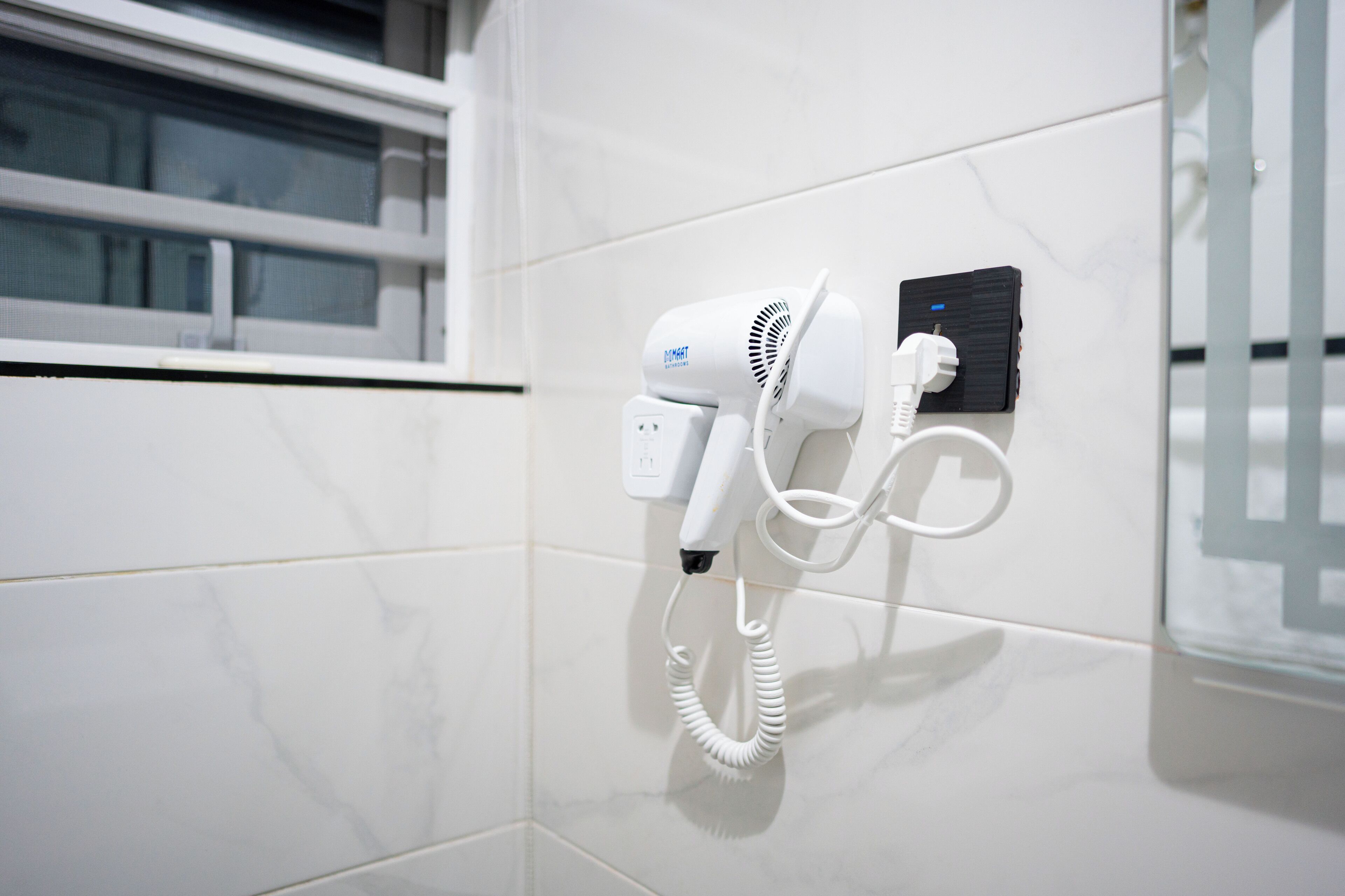 Un sèche-cheveux fixé sur un mur recouvert de carreaux blancs, avec une fenêtre au-dessus.