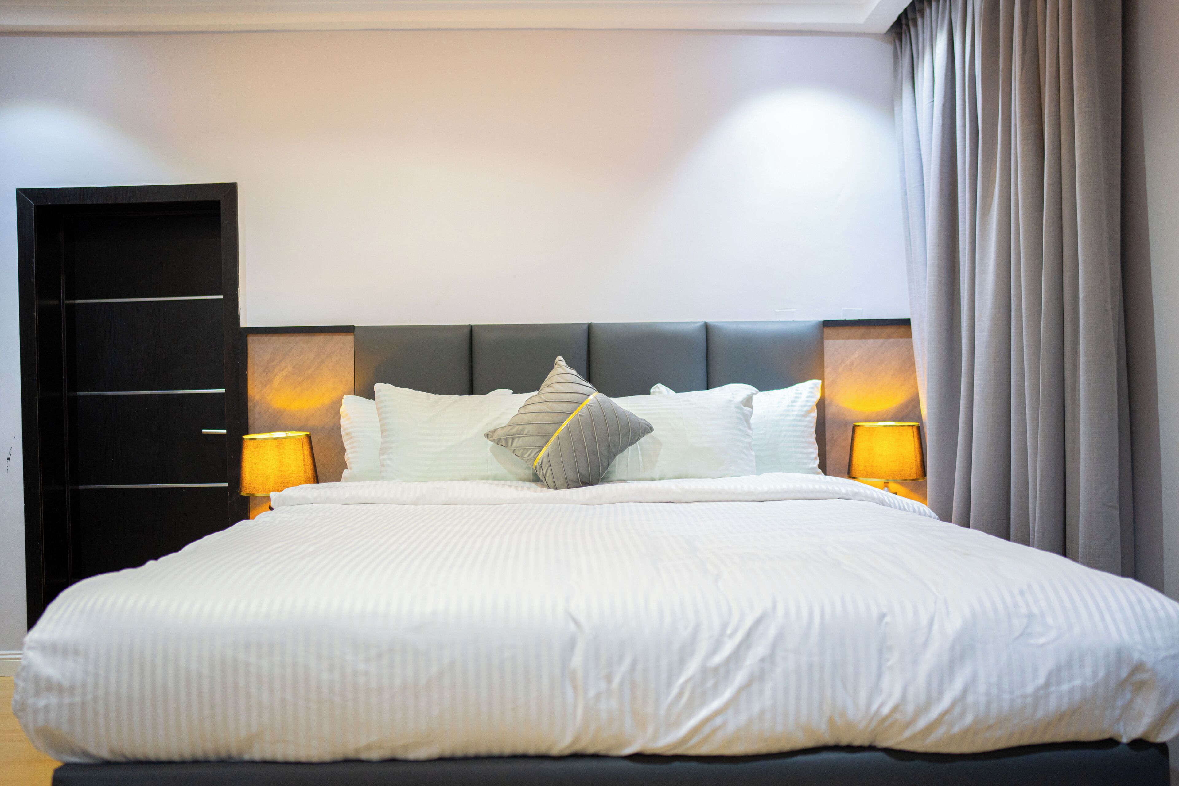 A neatly made bed with white bedding and multiple pillows, two bedside lamps, and a dark headboard in a hotel room.
