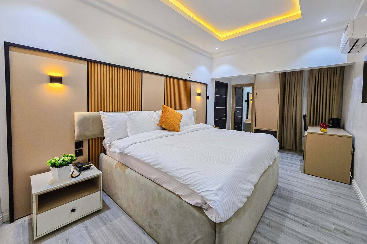 A modern hotel room with a large bed, bedside table, and a small desk with a chair.