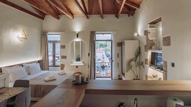 A modern hotel room with a wooden ceiling, a bed with white linens, a dining area with a wooden table, and a view of the street through a glass door. - Immerse yourself in Santa Lucia, a cozy, modern studio (Chania)