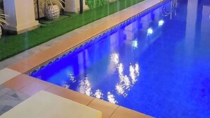 A swimming pool with a blue interior, surrounded by a tiled deck. There is a television mounted on a wall to the left, and a green wall with flowers in the background. - Morning Side Boutique Hotel and Suites  (Lagos)