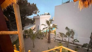 Vista desde un balcón con piscina, palmeras y un edificio blanco al fondo.