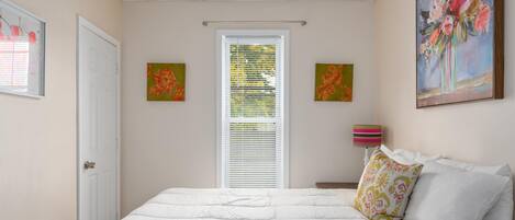 Un dormitorio con una cama, una ventana con persianas, una puerta y dos cuadros en la pared.
