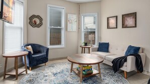 Una sala de estar con una mesa de centro redonda de madera, un sillón azul, un sofá blanco y una mesita auxiliar con una lámpara.