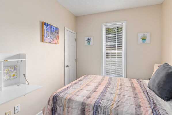 A bedroom with a bed, a door, a window with blinds, and wall-mounted artwork.