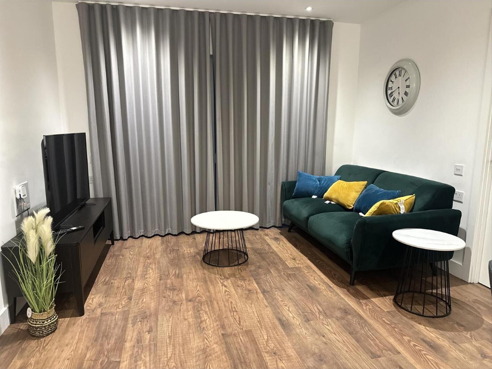 A living room with a green sofa, two white side tables, a black TV stand, and grey curtains.