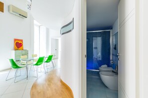 A modern interior with a dining area featuring a glass table and green chairs, a visible air conditioning unit, and a bathroom with a blue-tiled shower and a white bathtub. - Design Studio Duplex at National Museum (Naples)