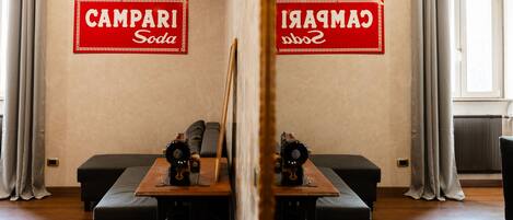 A room with a wooden table, two black sofas, and a mirror reflecting the room. There are two red signs on the wall reading "CAMPARI Soda."