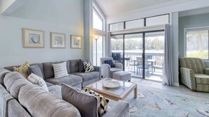 A modern living room with a grey sectional sofa, a glass coffee table, and a view of the outdoors through large sliding glass doors.