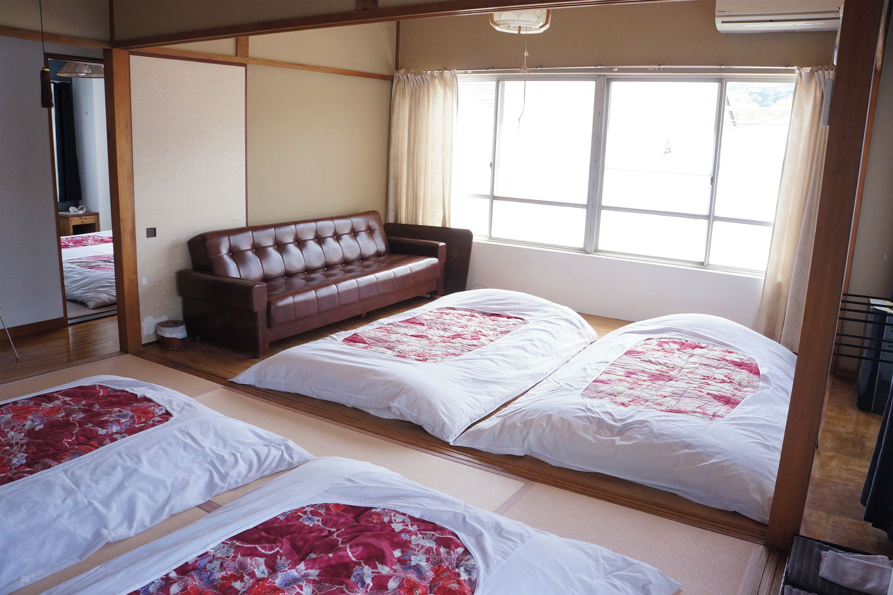 A traditional Japanese-style room with tatami flooring, a low wooden bed with a red and white floral bedspread, a brown sofa, and a large window with curtains.