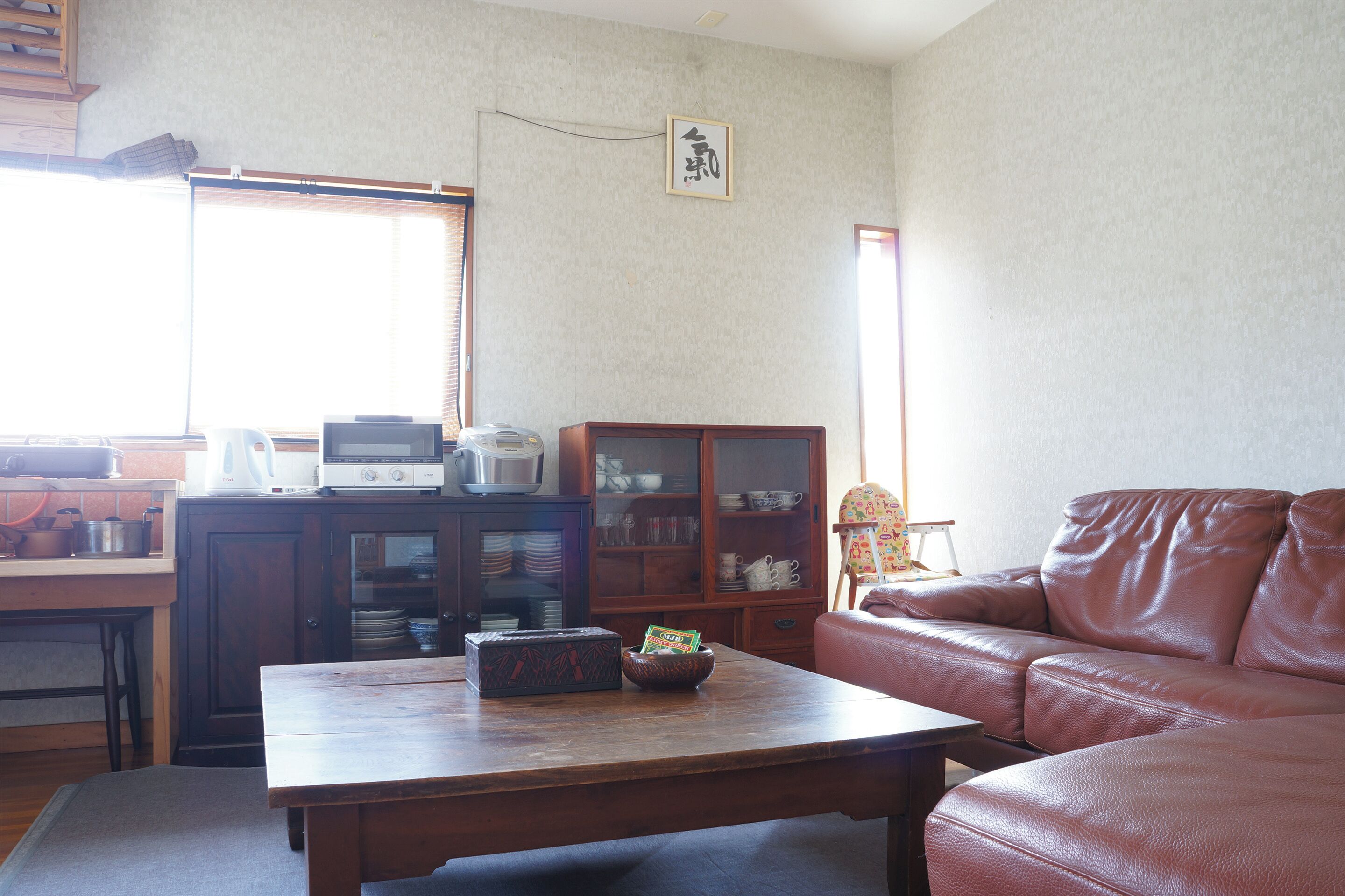 Una sala de estar con una mesa de centro de madera, un sofá de cuero marrón, un armario con vajilla y una ventana con persianas.