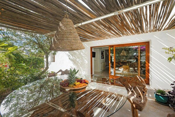 A modern outdoor dining area with a wooden table, a woven hanging light fixture, and a glass-top table reflecting the surrounding greenery.