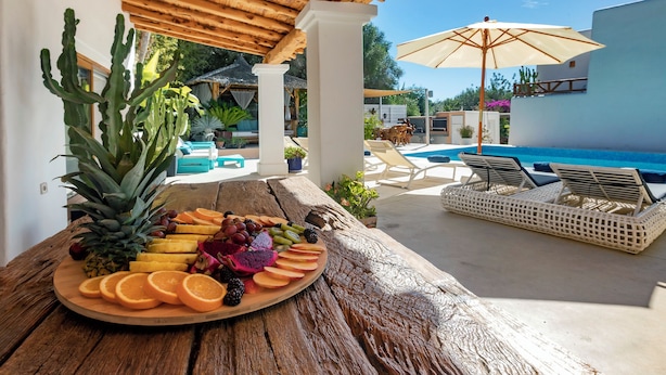 A wooden table with a fruit platter, a pool, lounge chairs, and a covered area with columns. - Villa near Ibiza Town, sleeps 13 (Ibiza)