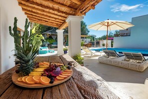 A wooden table with a fruit platter, a pool, lounge chairs, and a covered area with columns.
