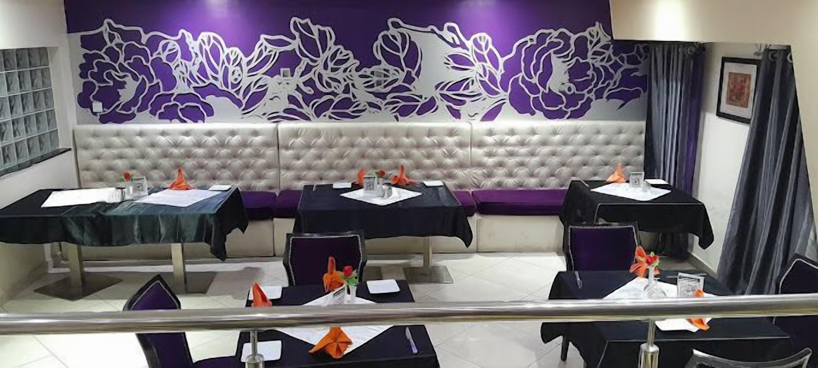 A dining area with a high ceiling, a decorative wall with floral patterns, and a cushioned bench. Tables are set with folded napkins and orange napkin rings.