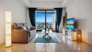 A modern living room with a sofa, a glass coffee table, a TV, and a view of the ocean through large windows.