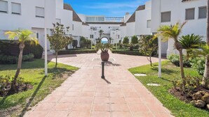 A courtyard with a central pathway, surrounded by white buildings with tiled roofs, palm trees, and a large potted plant.