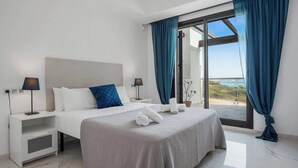 A modern hotel room with a large bed, two bedside tables, and a view of the beach through a sliding glass door.