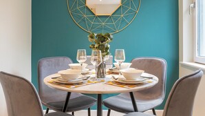A dining area with a round table set for four, a vase with flowers, and a geometric mirror on the teal wall.