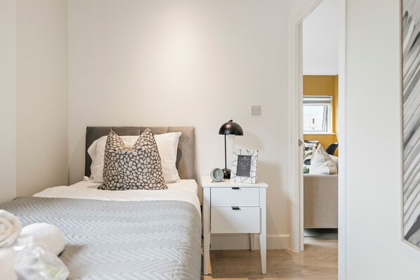 A neatly arranged bedroom with a bed, bedside table, lamp, and a view into another room.