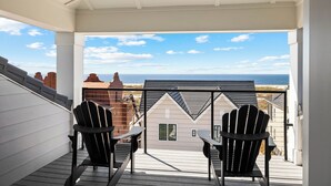 A balcony with two Adirondack chairs, a view of the ocean, and a building with a sloped roof in the background.