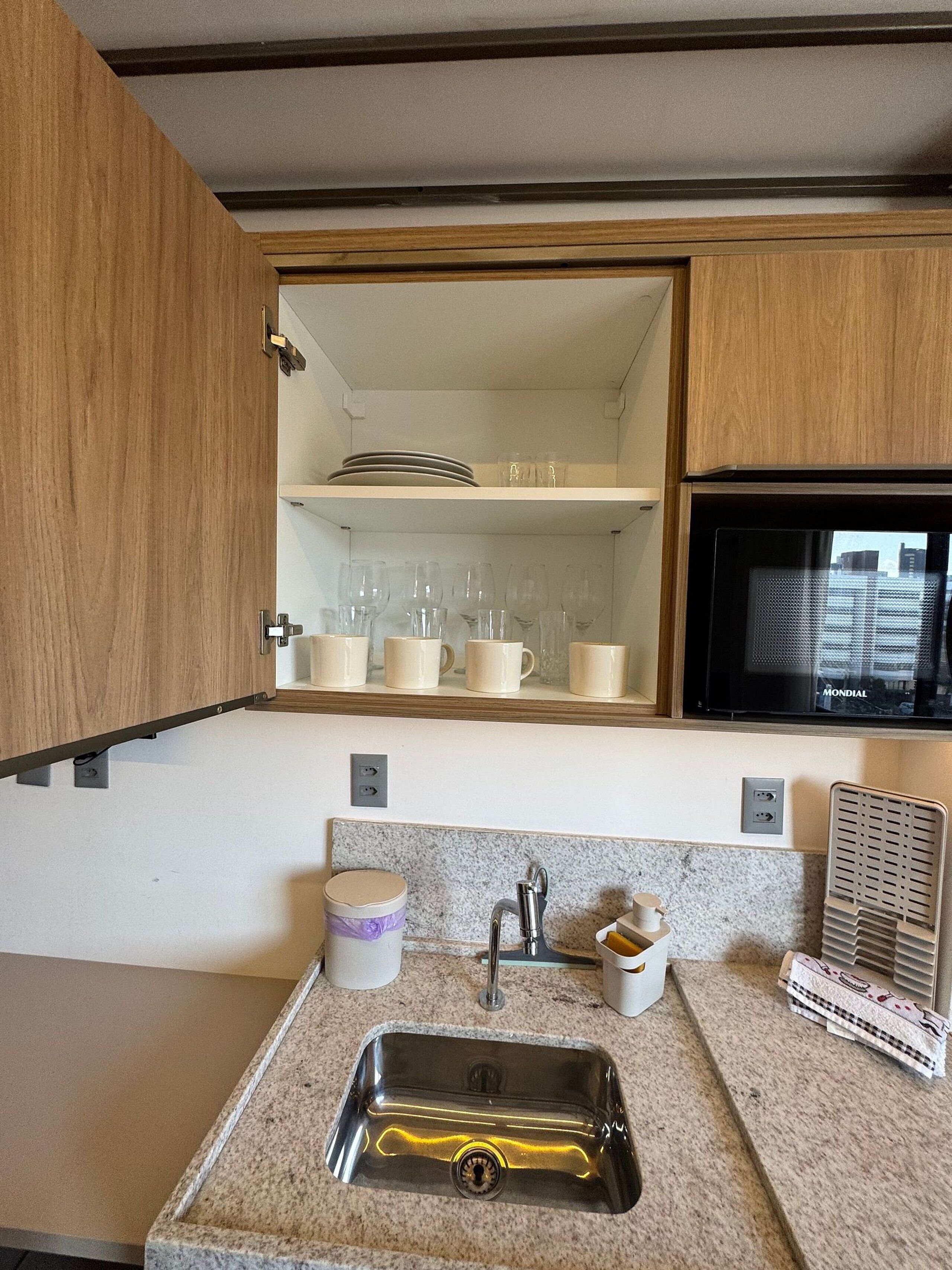 A kitchen with a stainless steel sink, wooden cabinets, a microwave, and a shelf with dishes and glasses.