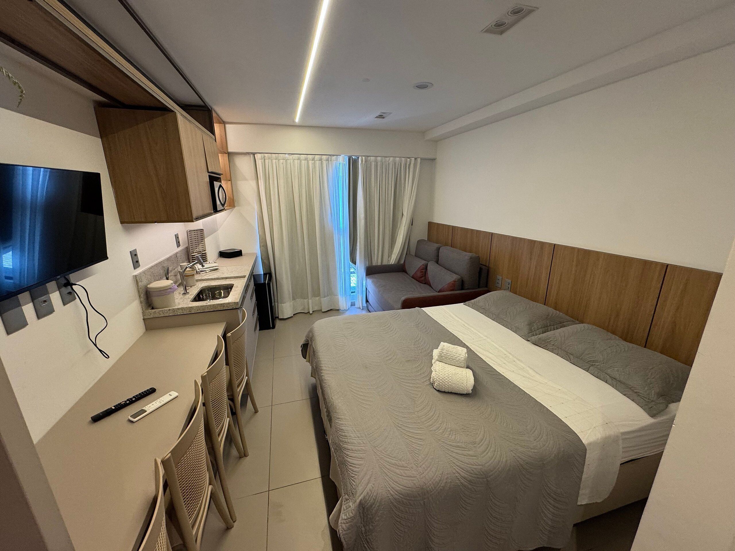 A hotel room with a bed, a desk with a TV, a small kitchenette, and a window with curtains.