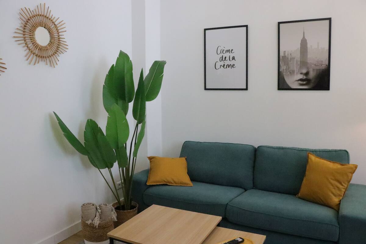 Un sofá color aguamarina con almohadones mostaza, una mesa de centro de madera, una planta verde grande y dos cuadros en la pared.