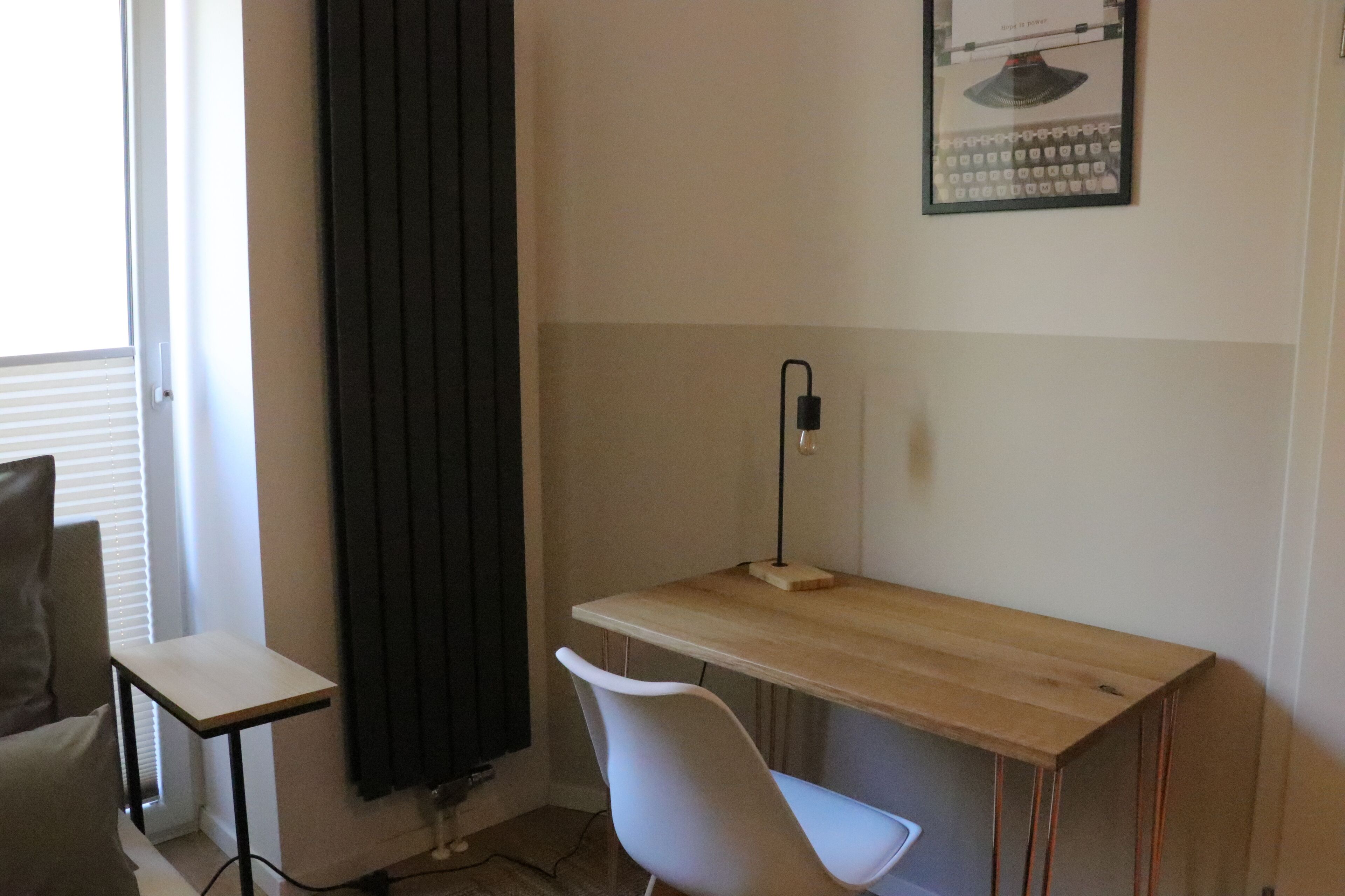 Een kamer met een houten bureau, een witte stoel, een zwart gordijn, een tafeltje en een poster aan de muur.