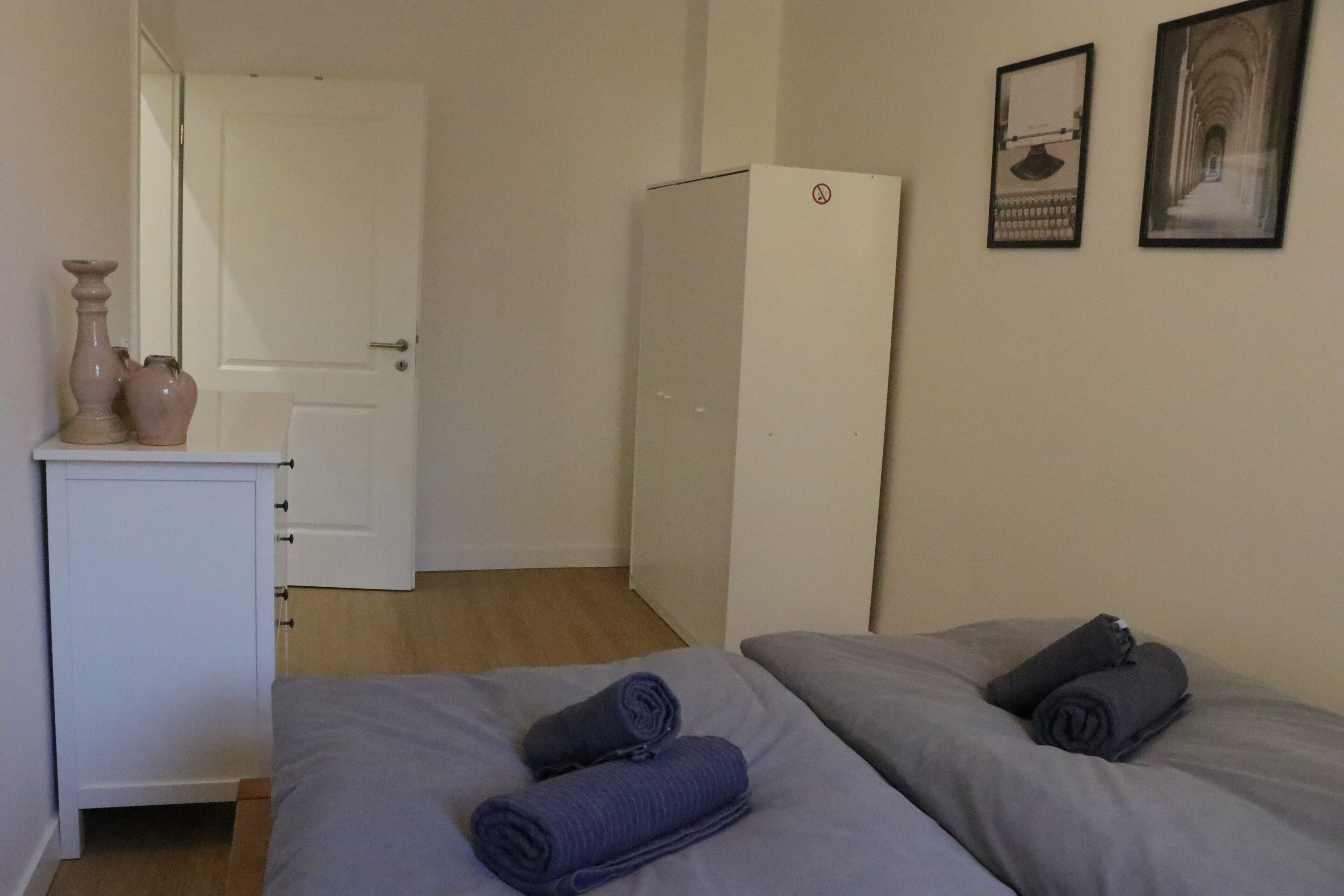 Ein Schlafzimmer mit zwei Betten, einem Kleiderschrank, einem Schreibtisch mit Spiegel sowie zwei gerahmten Bildern an der Wand.