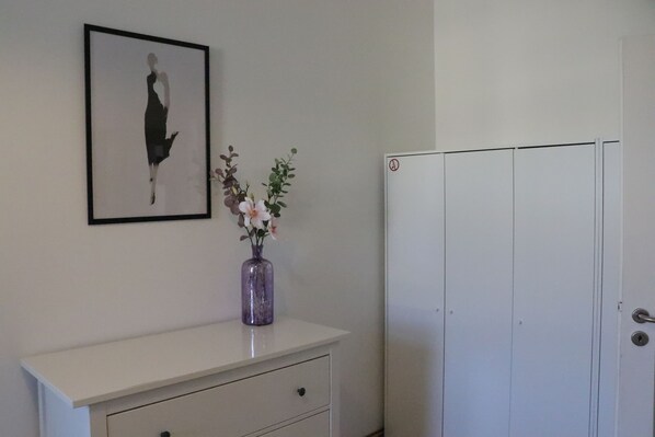 Un aparador blanco con una obra de arte enmarcada, un florero con flores y una puerta blanca.