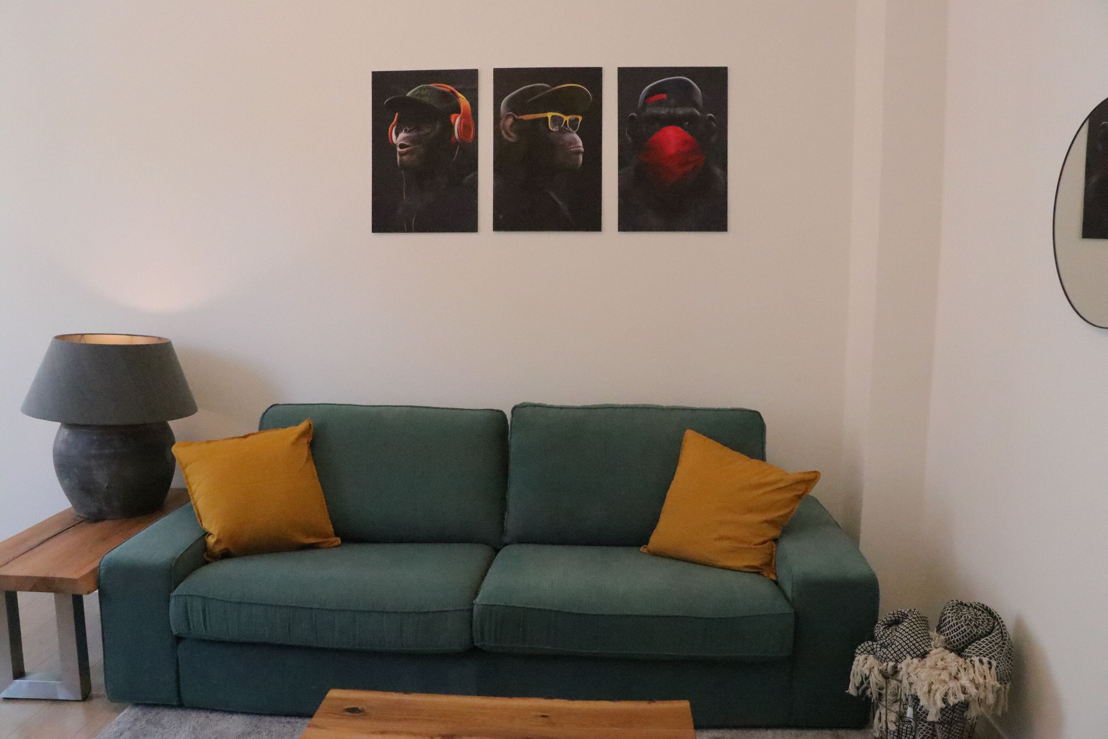 Een groene bank met twee gele kussens, een lamp en drie kunstwerken in lijsten aan de muur.