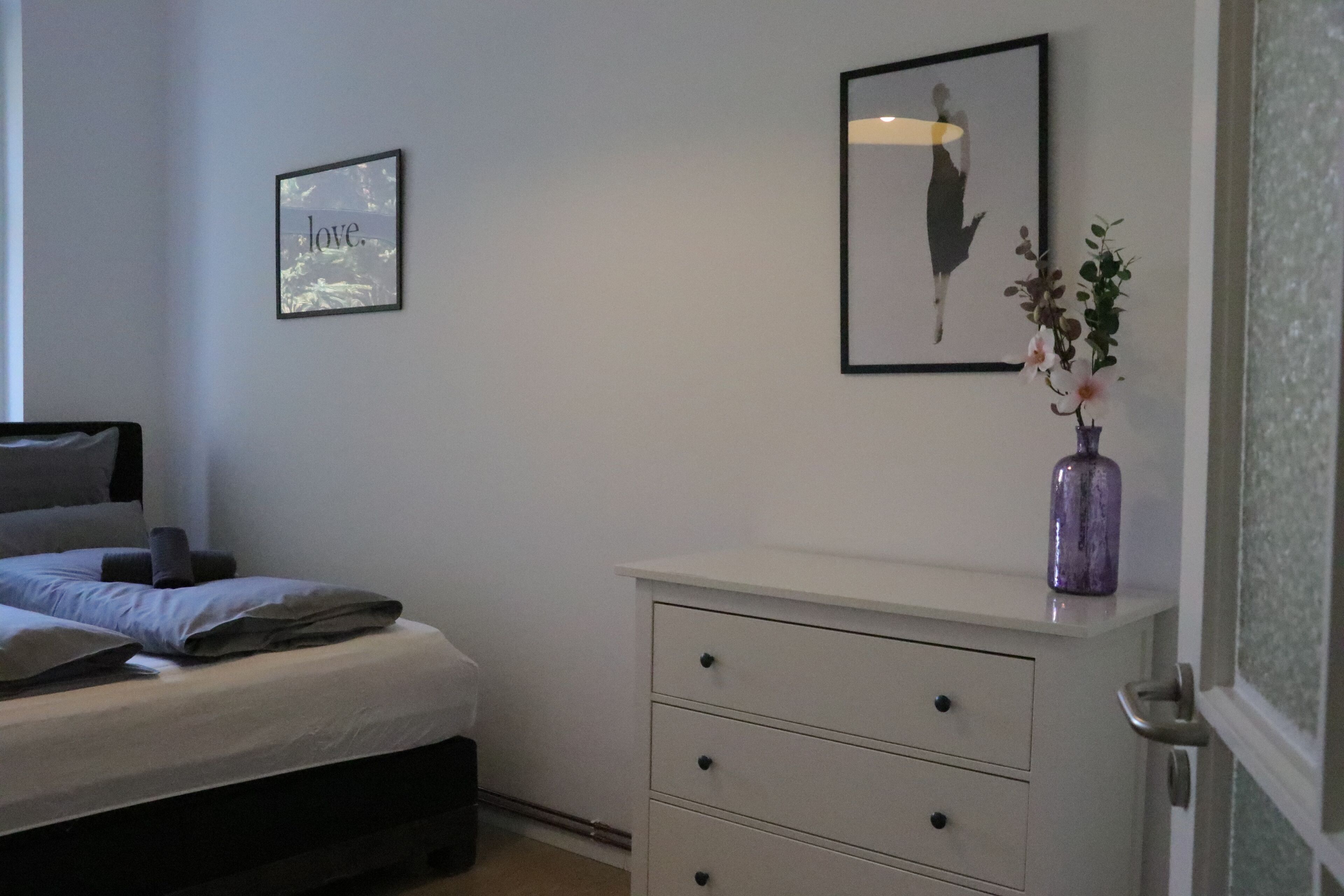 A bedroom with a bed, a dresser, a framed picture, and a vase with flowers.