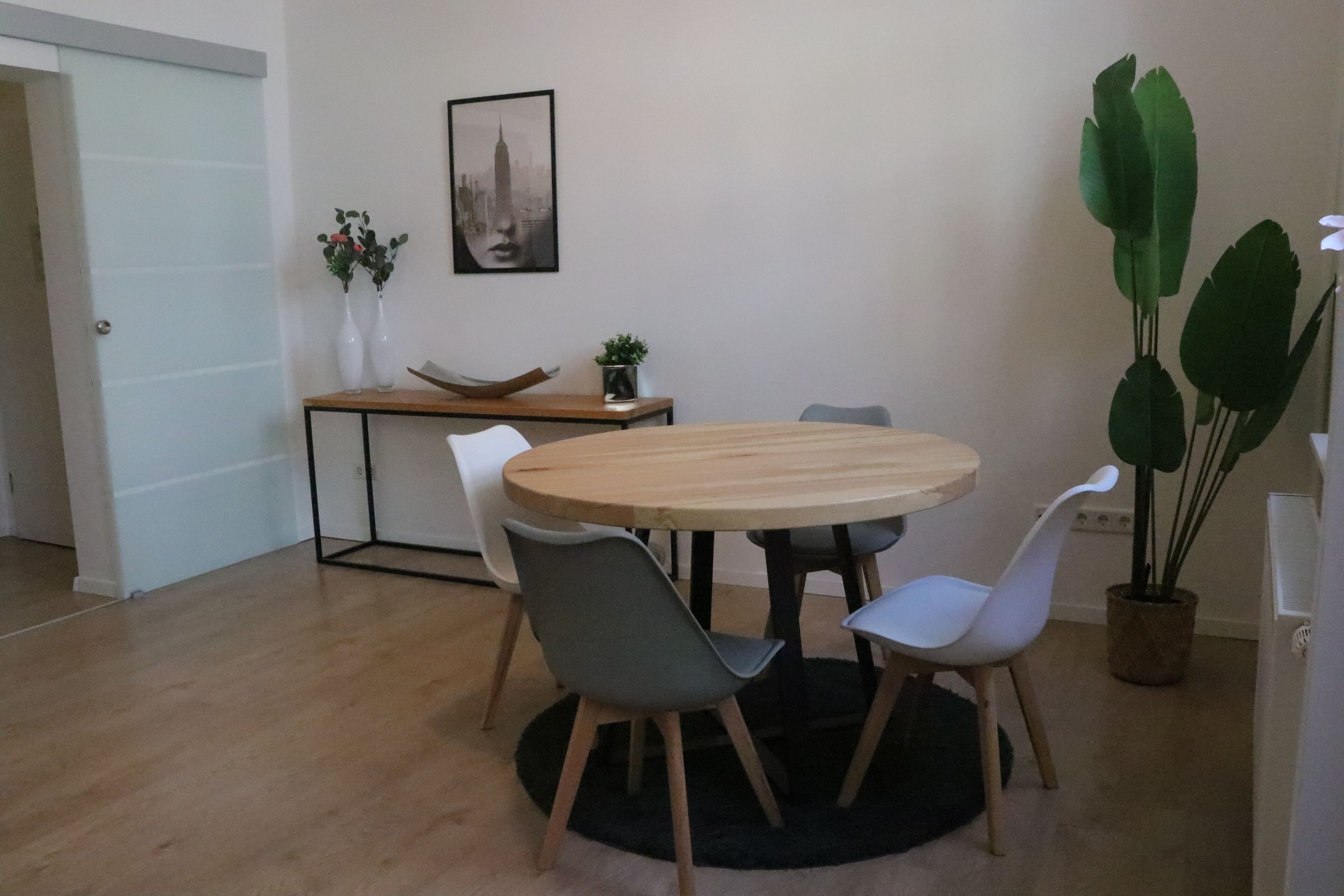 A dining area with a round table, four chairs, a small side table with a plant, and a framed picture on the wall.