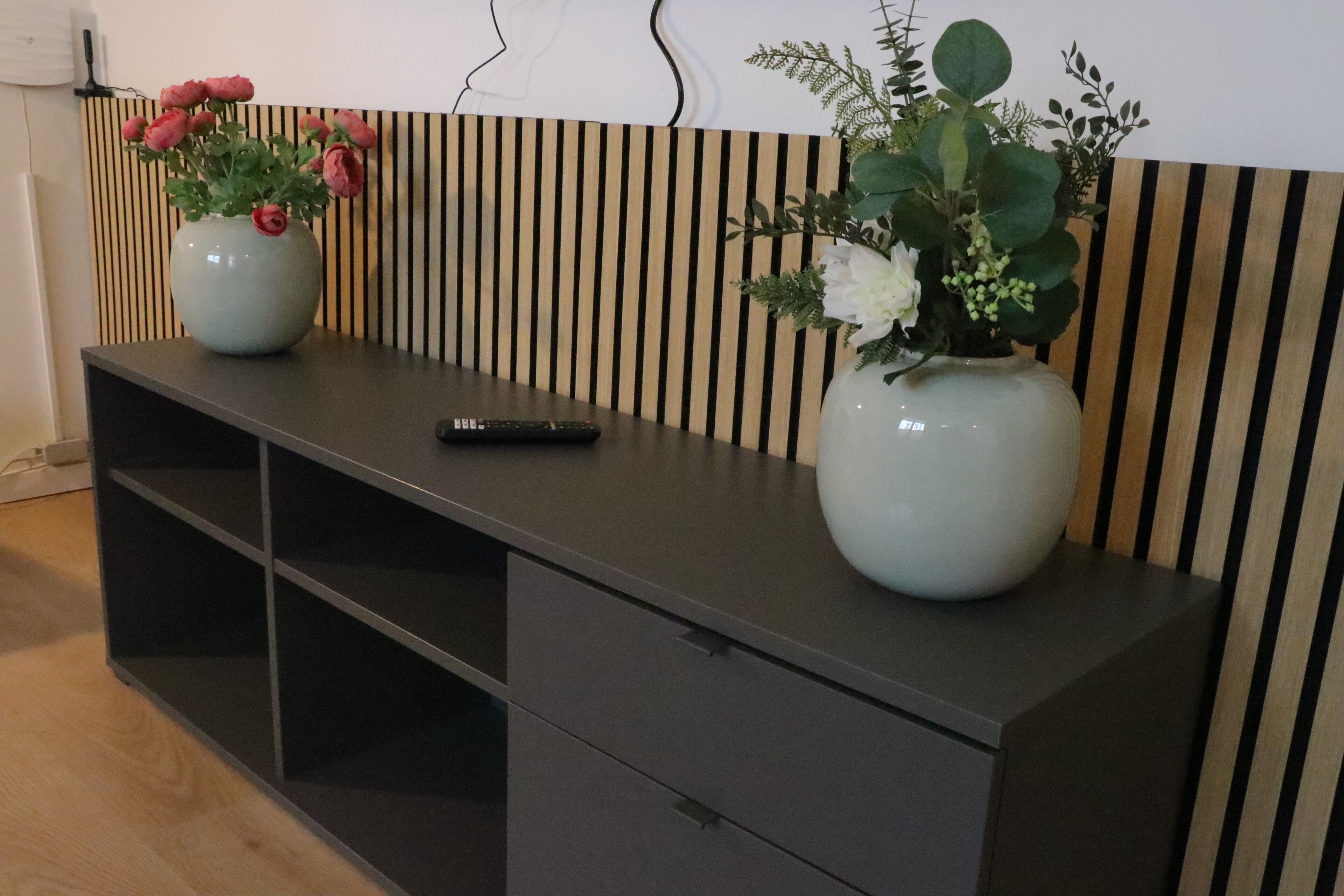 Ein modernes Zimmer mit einem dunkelgrauen Konsolentisch, zwei Vasen mit Blumen, einer Fernbedienung und einer gestreiften Wand.