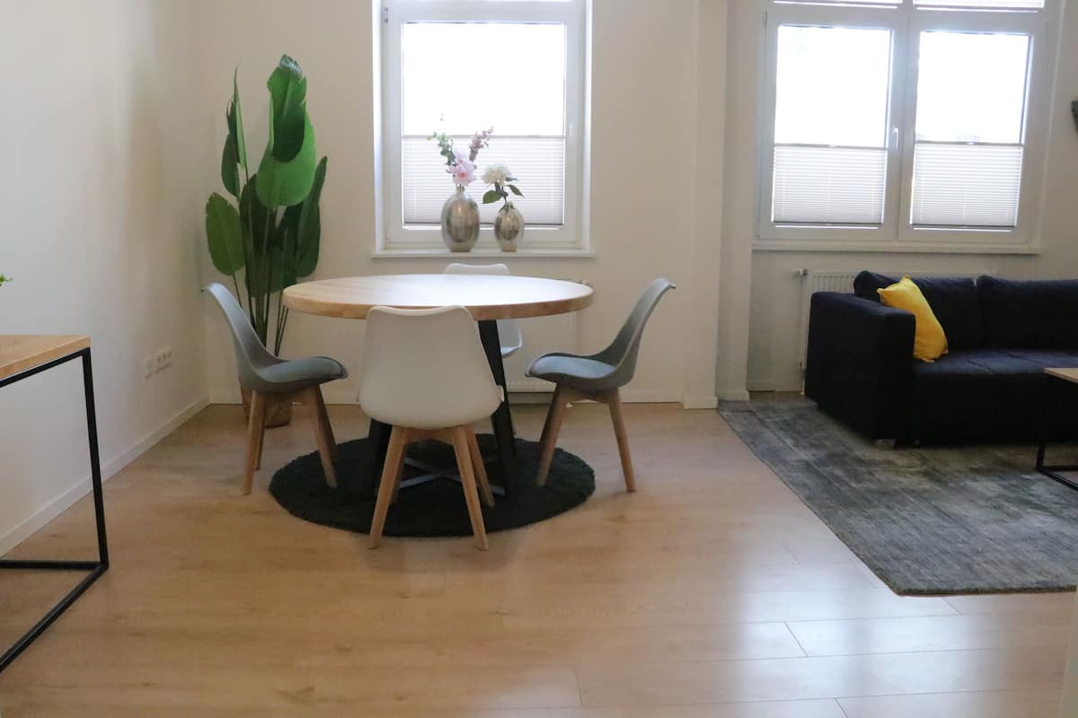 Un comedor con una mesa redonda y cuatro sillas, una planta en maceta, una ventana con persianas y un sofá de color oscuro con un cojín amarillo.