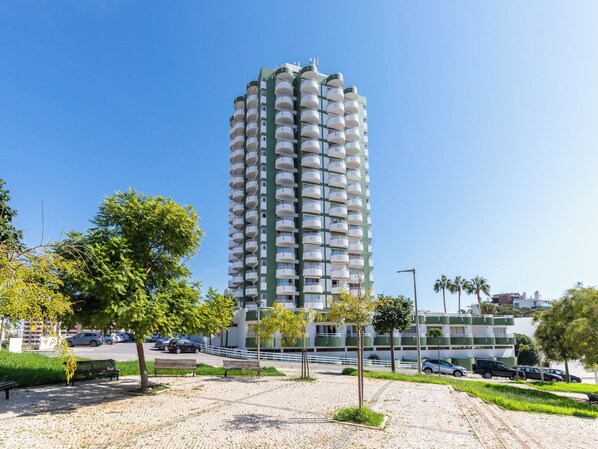 A high-rise building with multiple balconies, surrounded by a park with benches and trees.