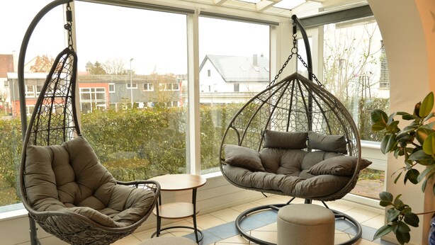 A room with two hanging chairs and a small table, large windows with a view of buildings and trees, and a potted plant on the right. - Vacation apartment Wintersun (Bad Mergentheim)
