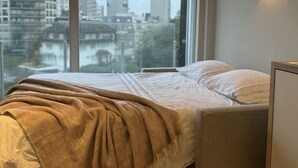 Una camera da letto con un'ampia finestra, un letto con coperta beige e un punto luce a parete.