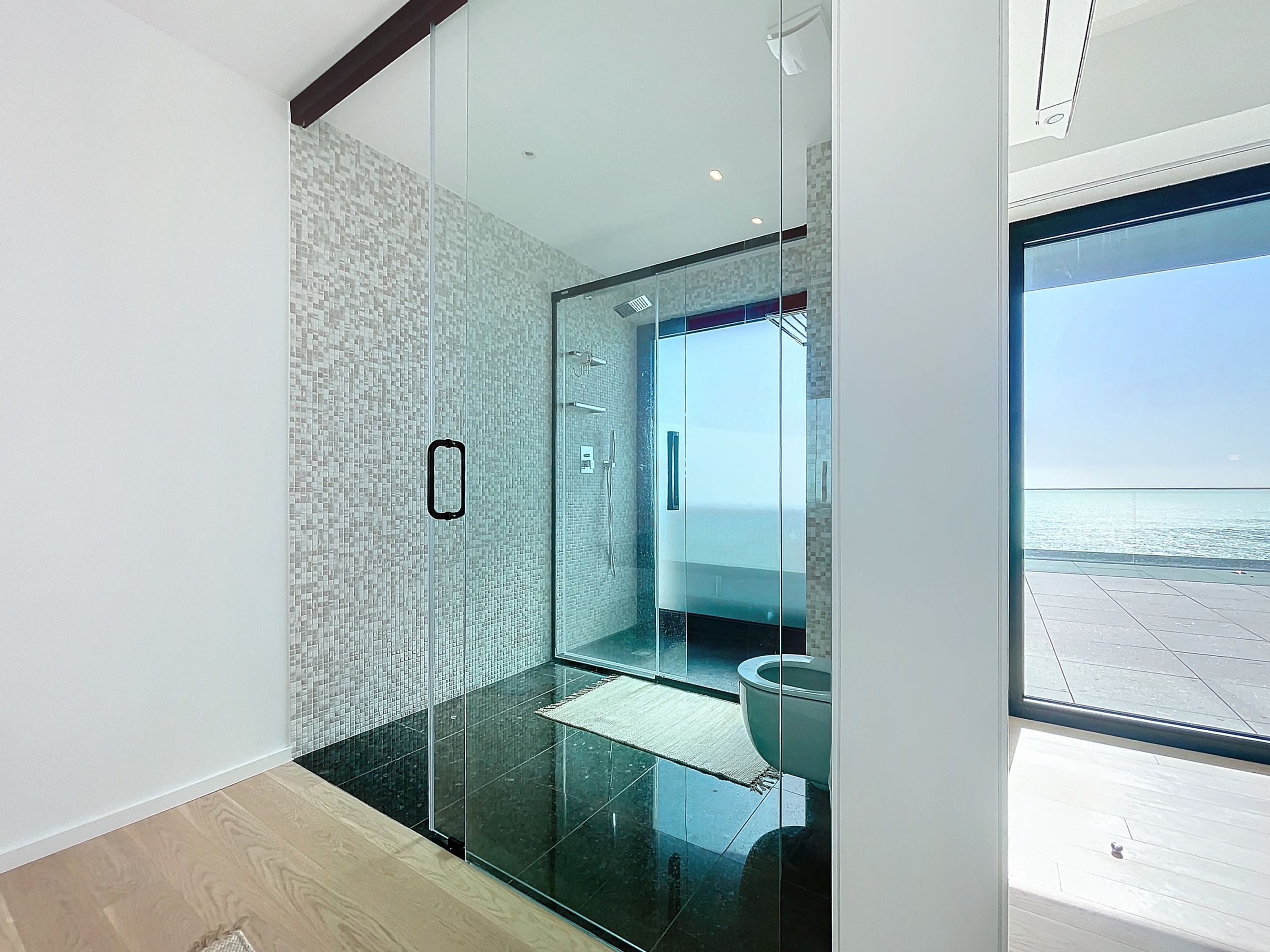 Ein modernes Badezimmer mit Duschkabine aus Glas, sichtbarer Toilette und Meerblick durch ein großes Fenster.