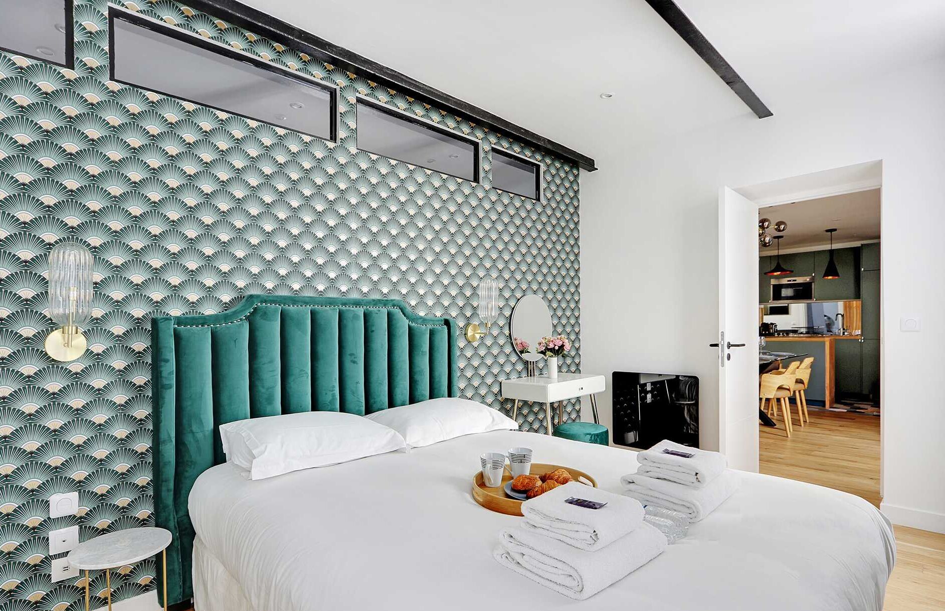 A bedroom with a teal upholstered headboard, a white bedspread, and a tray with croissants and cups.