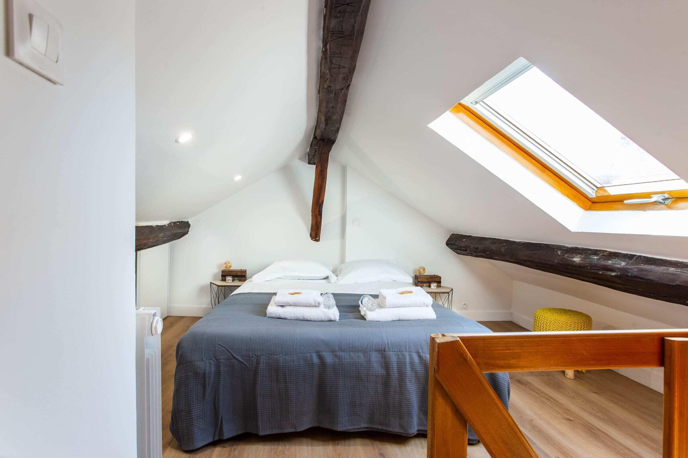Une chambre avec un plafond en pente, un puits de lumière, un lit avec du linge de maison blanc, un banc en bois et un petit pouf jaune.