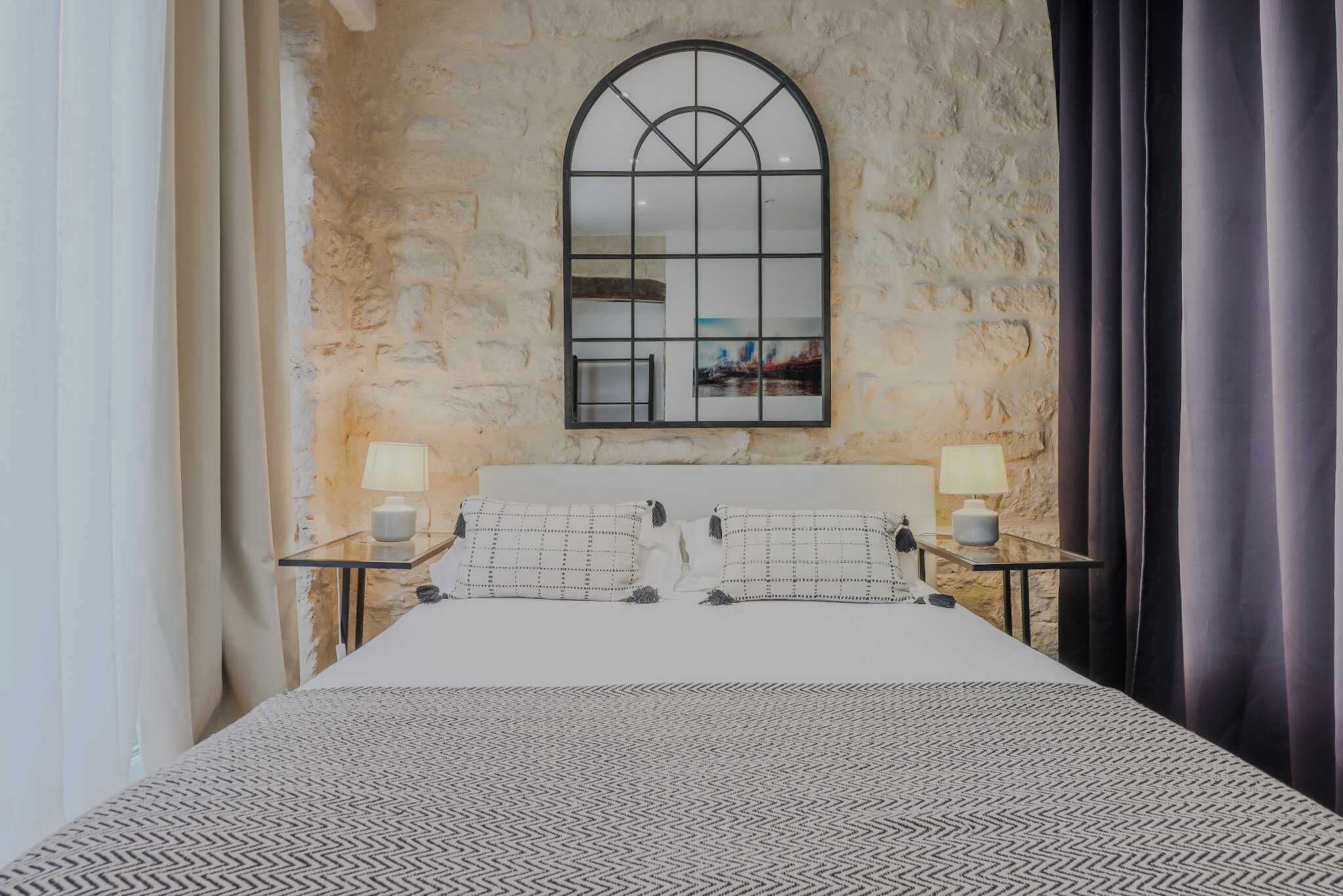A bedroom with a stone wall, a large arched mirror, a bed with patterned bedding, two bedside tables with lamps, and a view of a room with a painting.