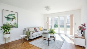 A modern living room with a grey sectional sofa, a round coffee table, and a large window with a view of a garden. - Luxurious 4 BH Home Near Willen Lake Ideal for Families & Business Stays! (Milton Keynes)