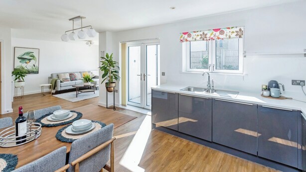 A modern kitchen with dark cabinets, a sink, and a window above it. There is a dining area with a table set for two, a sofa, and a plant. - Luxurious 4 BH Home Near Willen Lake Ideal for Families & Business Stays! (Milton Keynes)